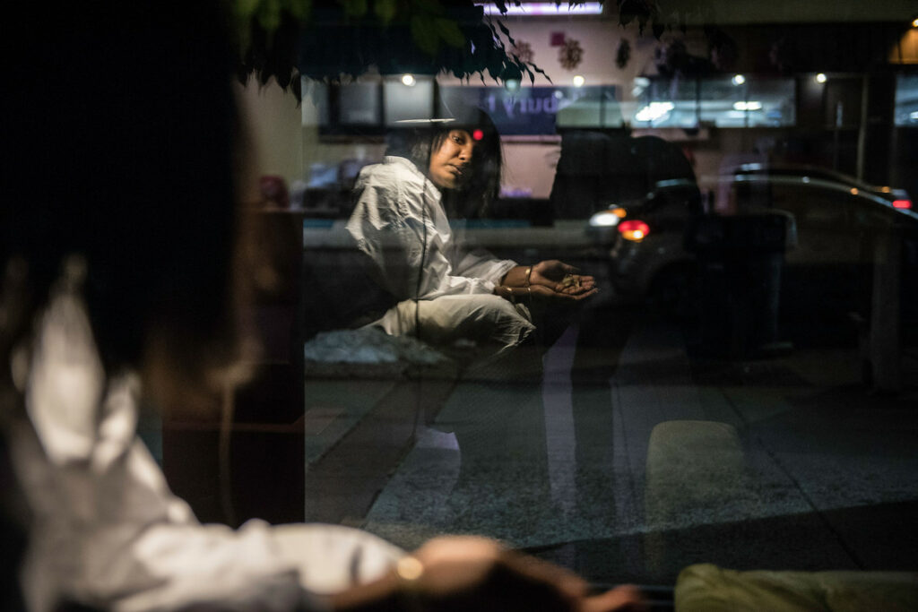 Photo of a brown woman looking into her own reflection with her hands reached out in the lobby of Pillsbury House + Theatre. Through the window of her reflection is a car passing by.