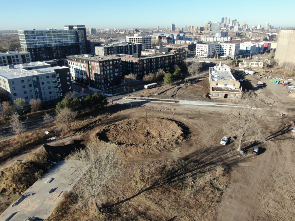 An aerial view of a neighborhood centered on a large empty space with a convex mound of exposed dirt