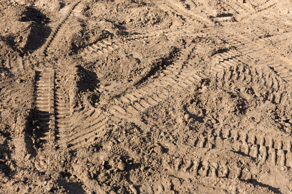 Close up image of loose light brown dirt, marked with tire treads and a building in the background