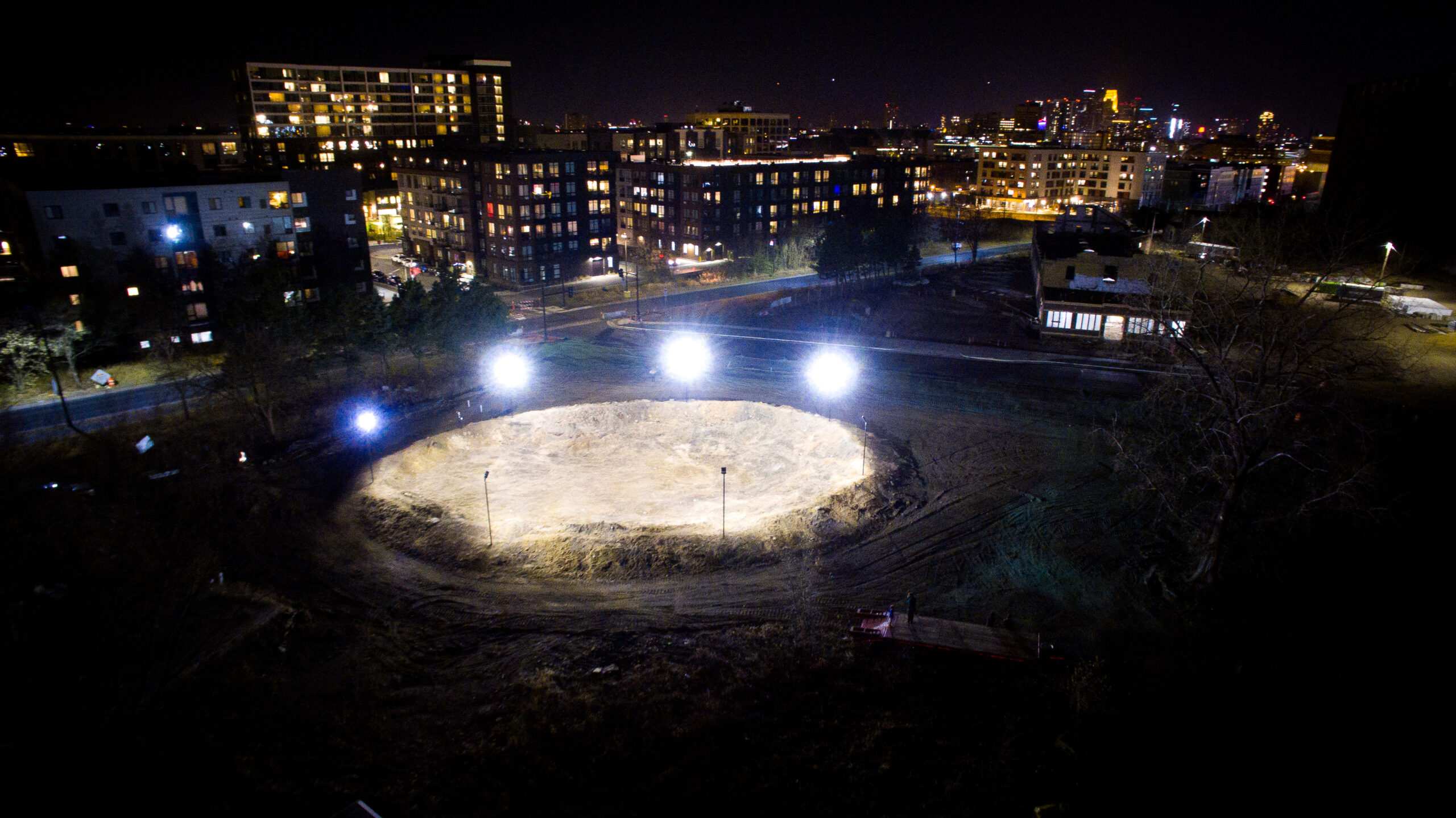 Aerial view of a convex mound of dirt lit with stadium lights at night, with apartment buildings in the background