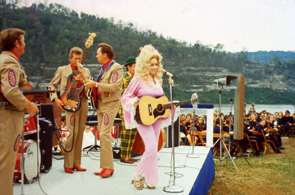 Dolly Parton stands on a stage in front of a microphone with a guitar, wearing a pink jumpsuit. Men in decorative brown suits are behind her on the stage by instruments, an audience is sitting in chairs behind them, and a body of water and green mountains are in the background.