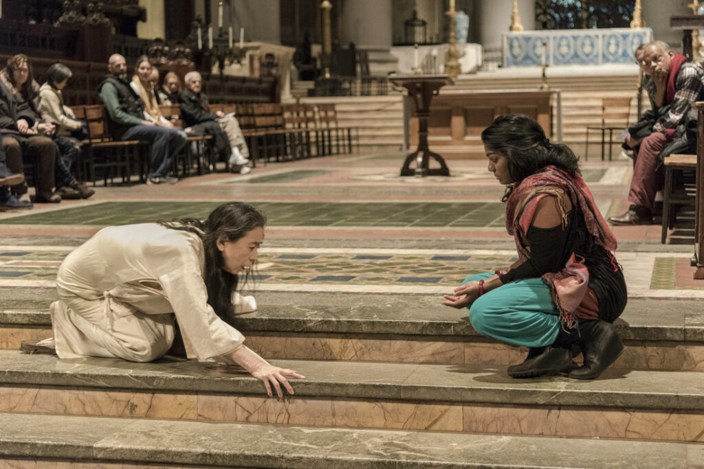 Photo of two women sitting on the stairs of a cathedral, one woman dark-skinned who is draped in a colored shawl reaching her hands out on her lap as she gazes at the other woman, who is draped white and crouching on the stairs. People are witnessing behind them.