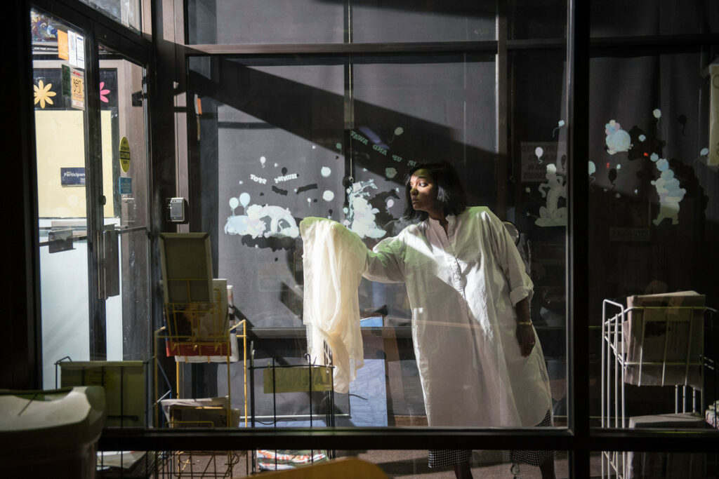 Photo of a brown woman extending a white fabric out from her body in the doorway of Pillsbury House + Theatre. A light shines on her eyes through the doorway.