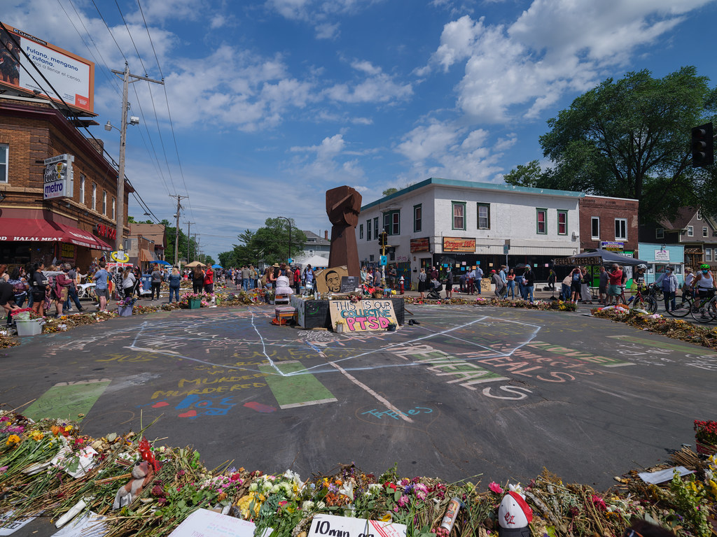 An intersection with a memorial for George Floyd at the center that has a large sculpture of a fist in the air. Signs and flowers are arranged in a circle around the memorial, and people can seen standing on the outside looking in