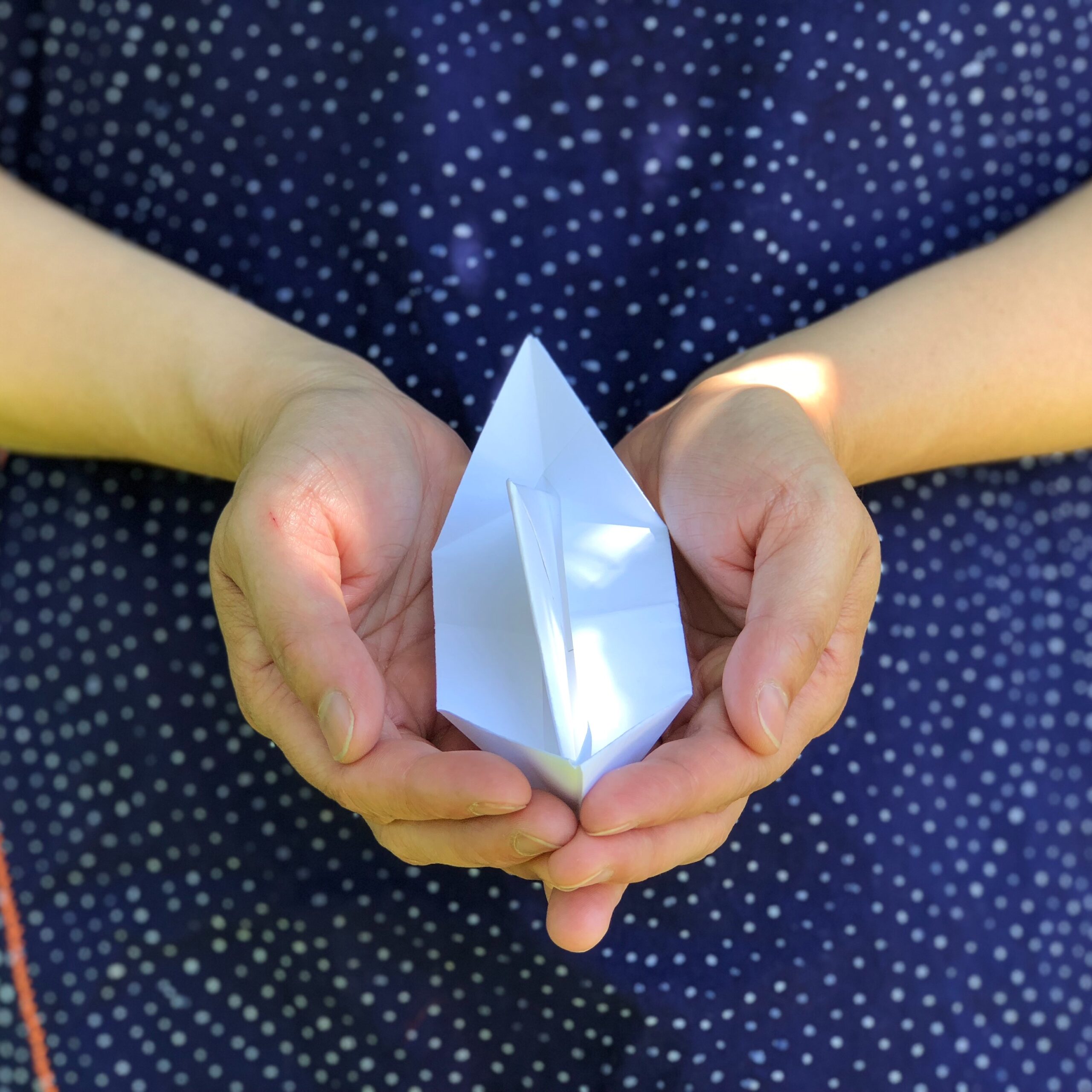 Two hands cup a white paper boat, against a background of blue fabric.