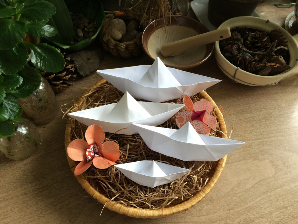 Four white paper boats are arranged in a wicker basket, surrounded by paper flowers, plants, jars, bowls, and pinecones.