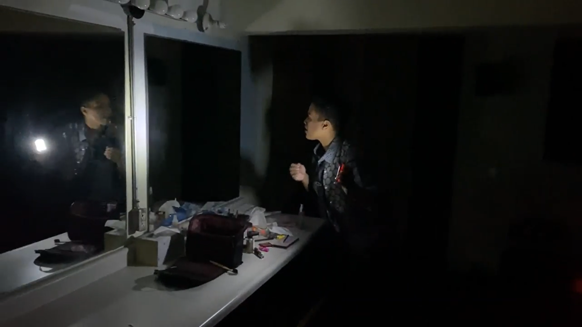 Mx examines themselves in a dark dressing room mirror holding their light.