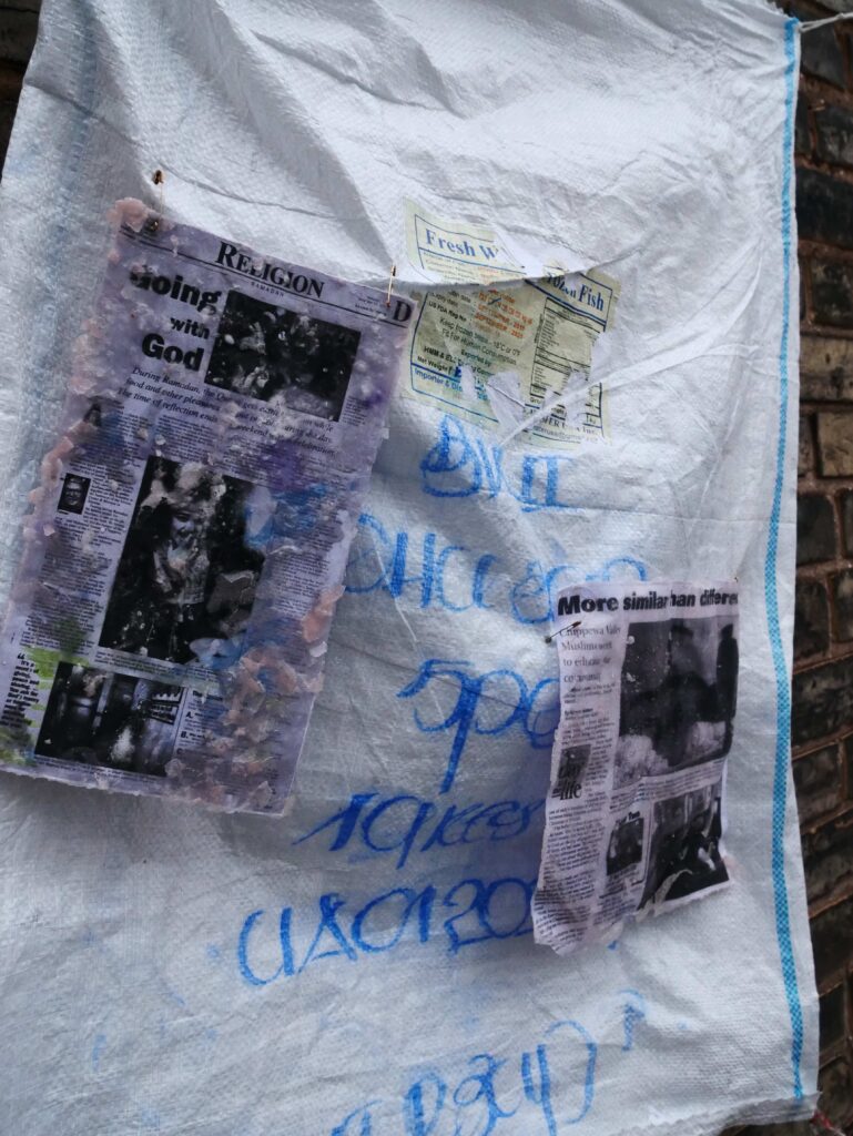 A white sack with blue illegible print on it is depicted with two water-worn news articles attached. one article reads "going with god" as its header.