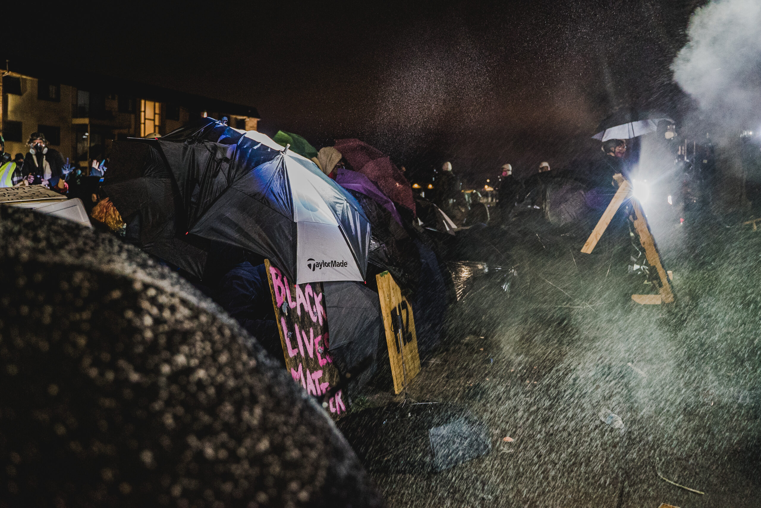 Clouds of smoke in front of a row of protesters' umbrellas, and a sign reading BLACK LIVES MATTER.