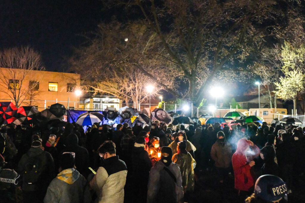 A crowd of protesters with masks, umbrellas, and candles, under a tree, street lights, and night sky.