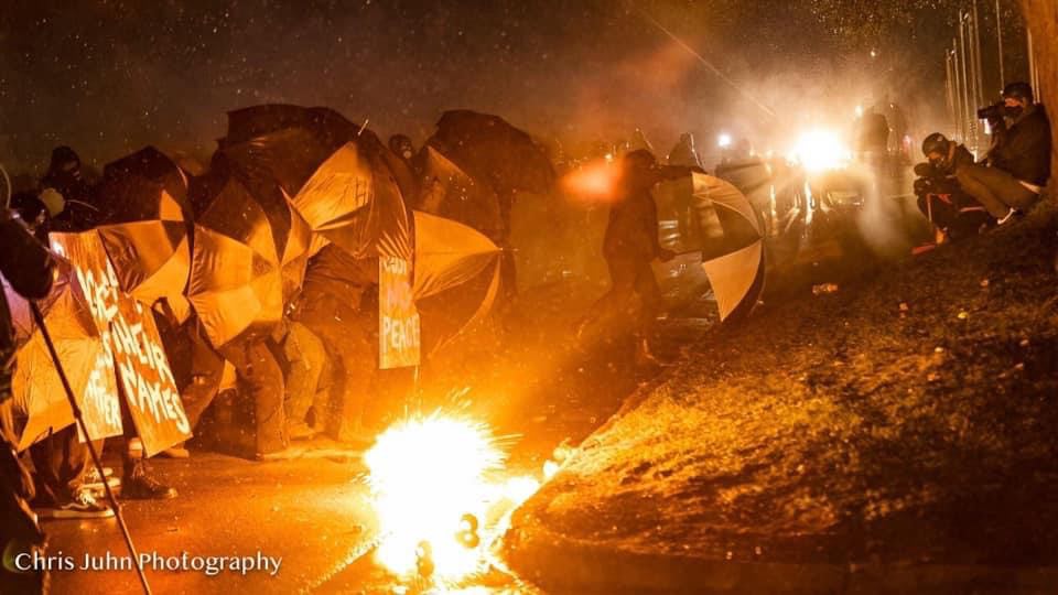 Protesters shield themselves with umbrellas as a flash bang explodes in front of them on the sidewalk.