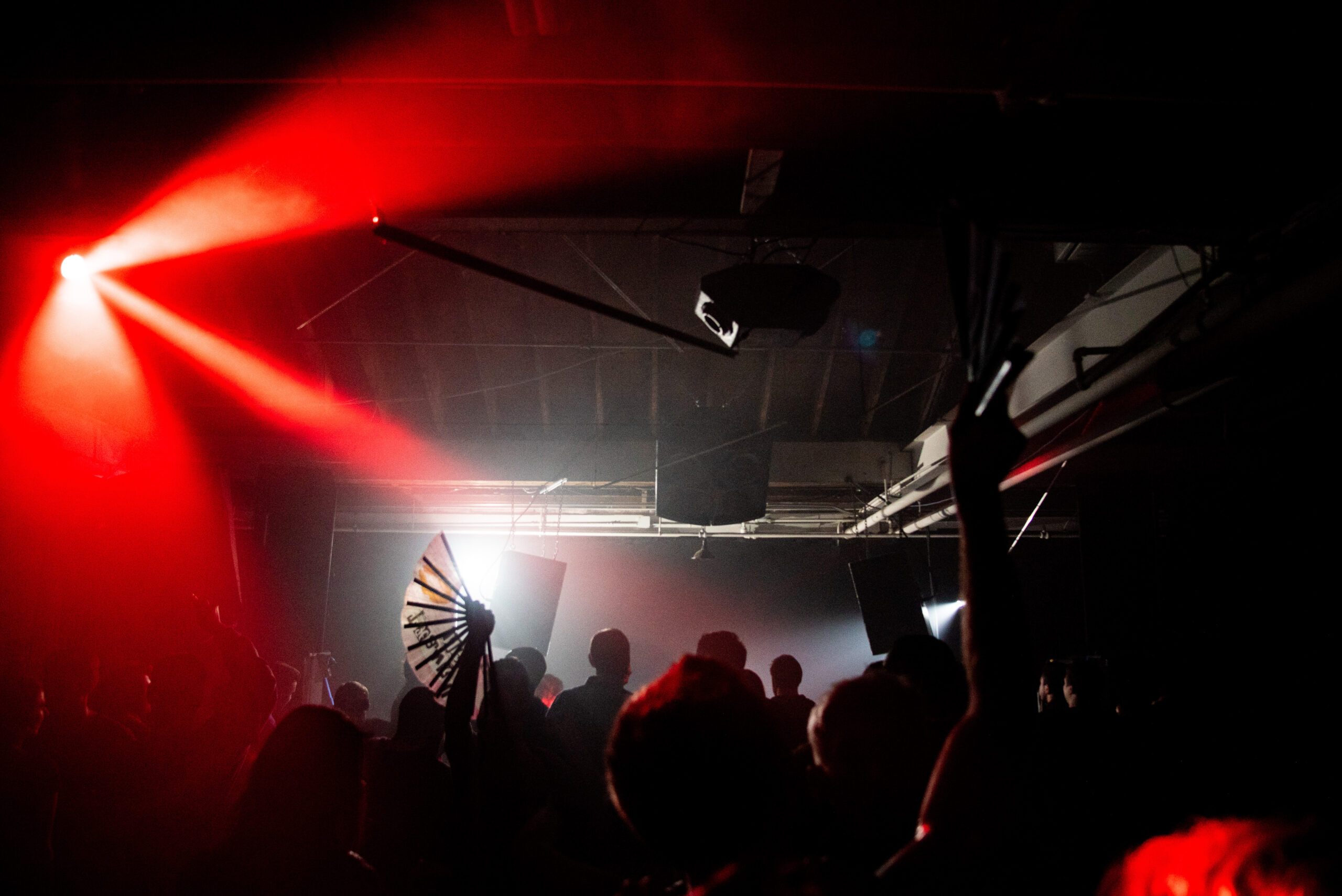 Three beams of red light over a crowd on a dark dance floor, and a white fan raised above the crowd.