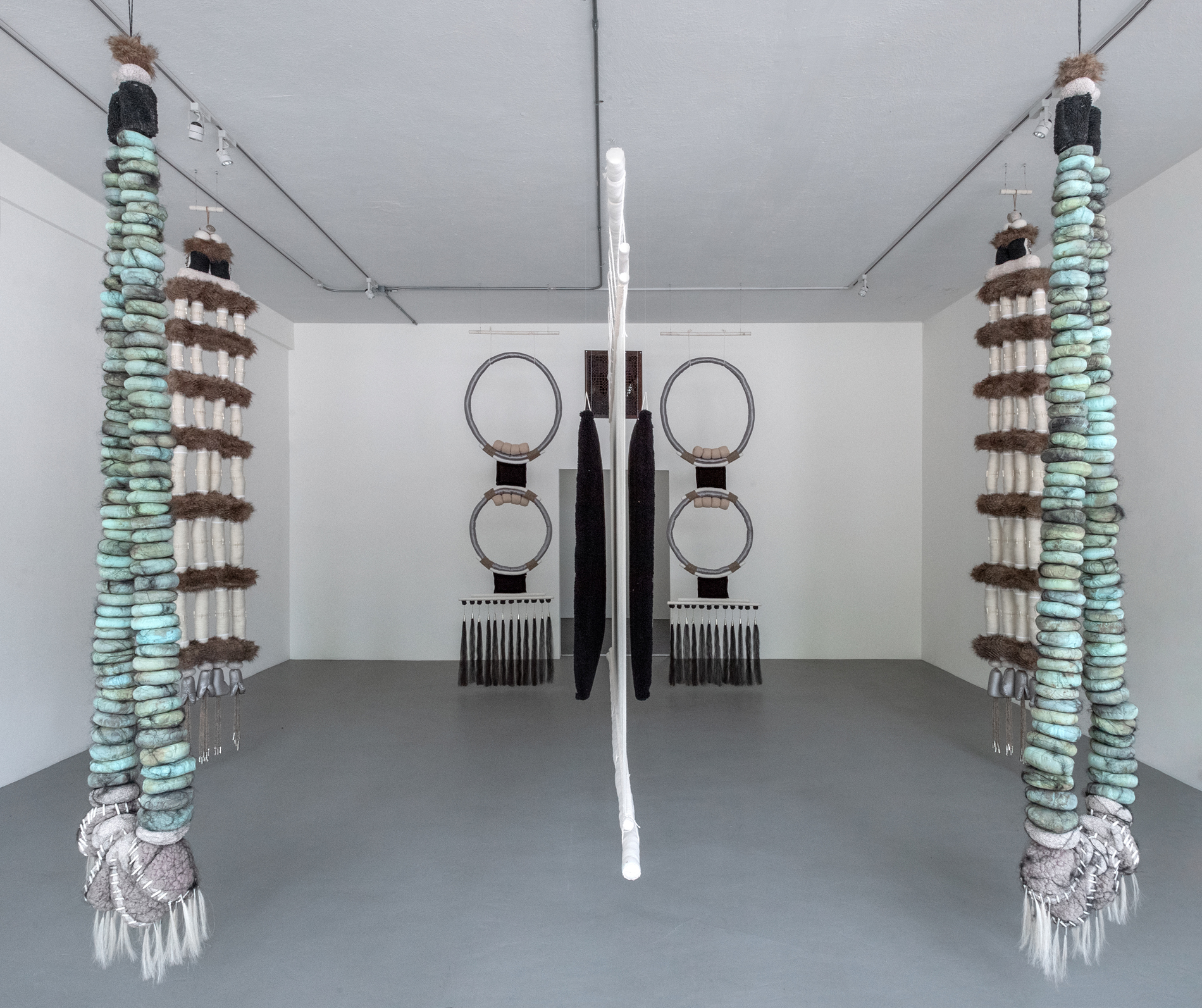 Installation view of jewelry-like fiber sculptures hanging in a gallery with white walls and gray floors.