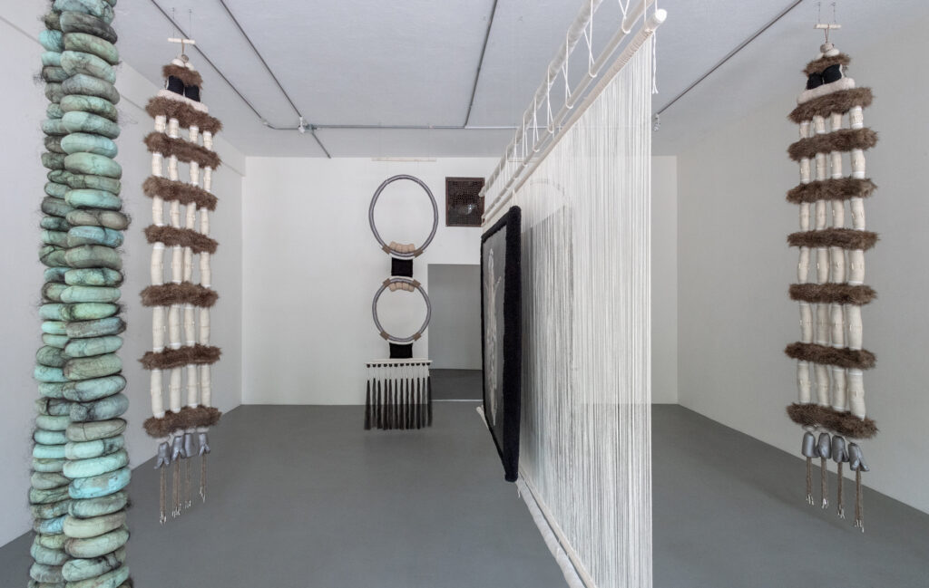 Installation view of jewelry-like fiber sculptures hanging in a gallery with white walls and gray floors.