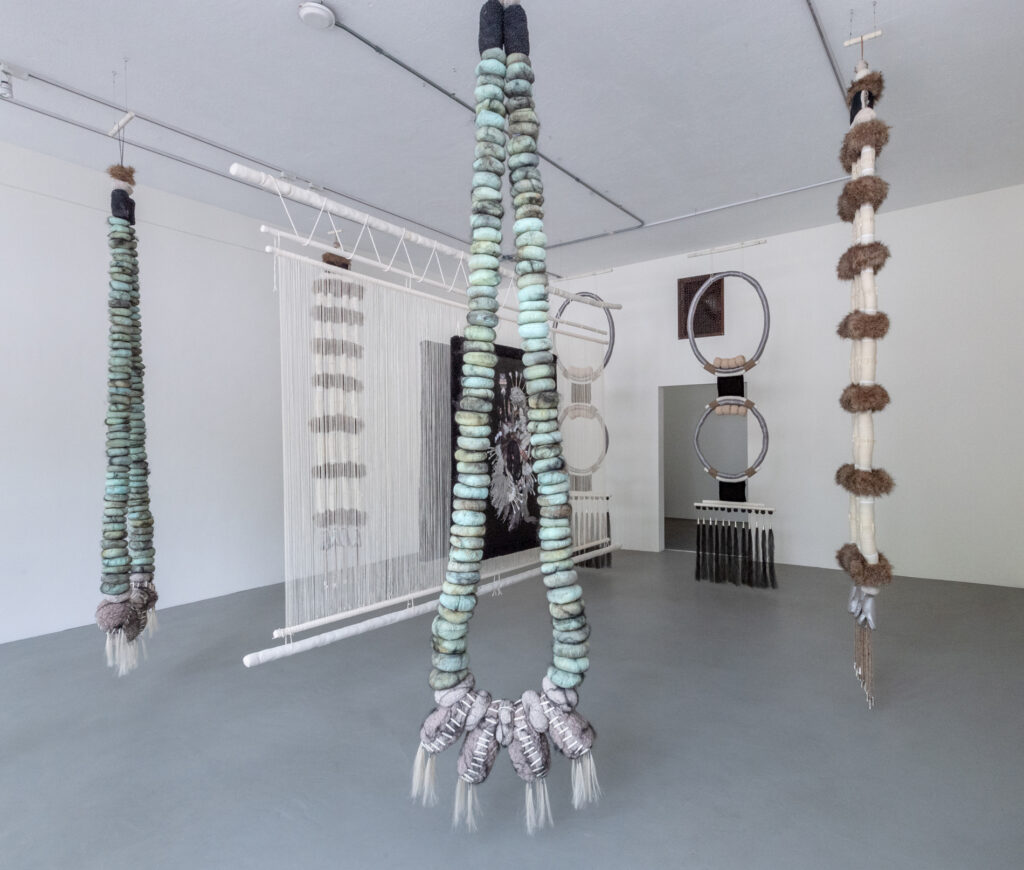 Installation view of jewelry-like fiber sculptures hanging in a gallery with white walls and gray floors.