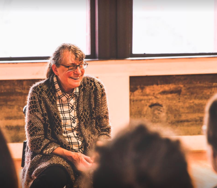 Person with gray ponytail, glasses, plaid shirt and gray sweater sits in front of a window.