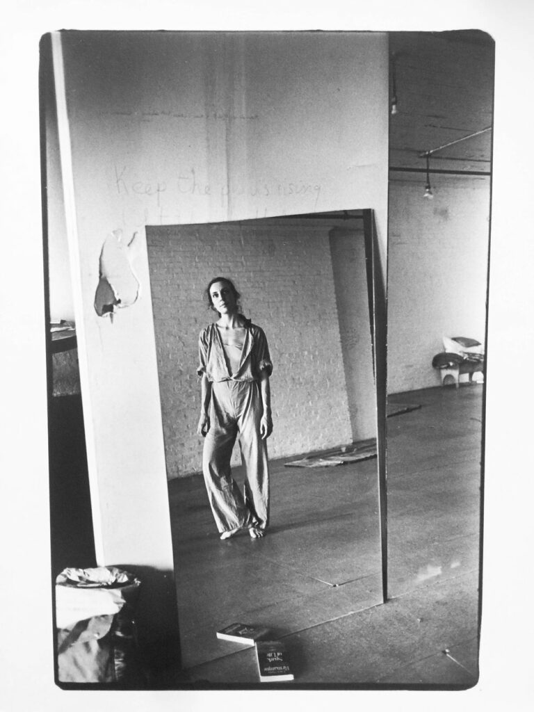 Black and white photo of Mary Overlie, looking into a mirror with heel raised off the ground.