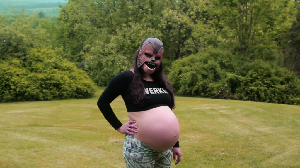 A person wearing a Chewbacca mask stands in a green field, exposing their large pregnant belly.