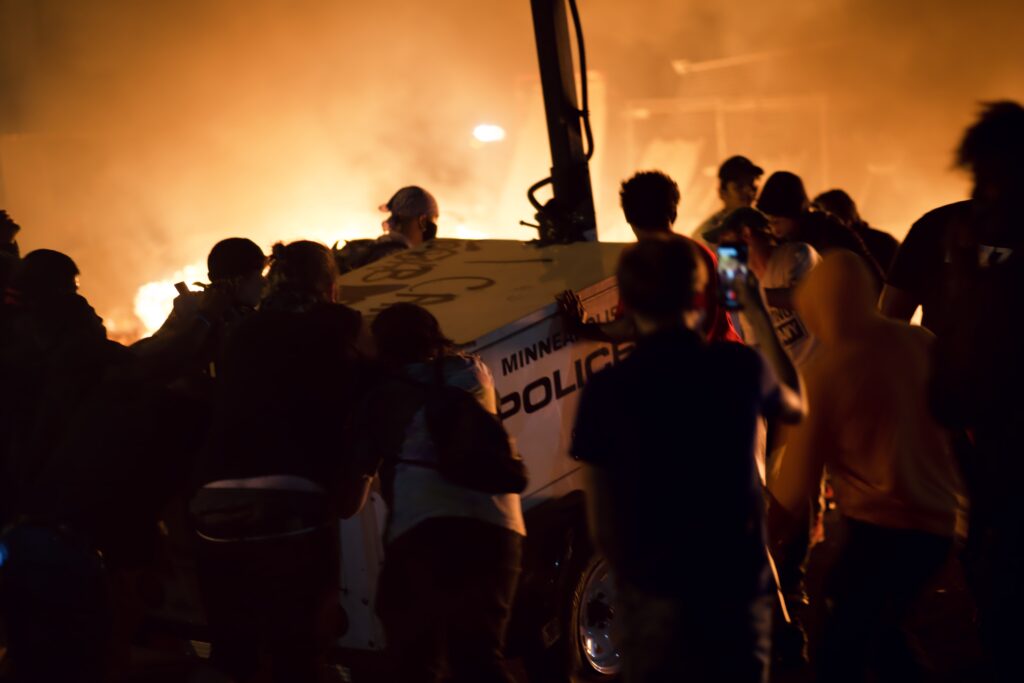 Crowd of people surrounds police mobile surveillance trailer, with fire glowing in background.