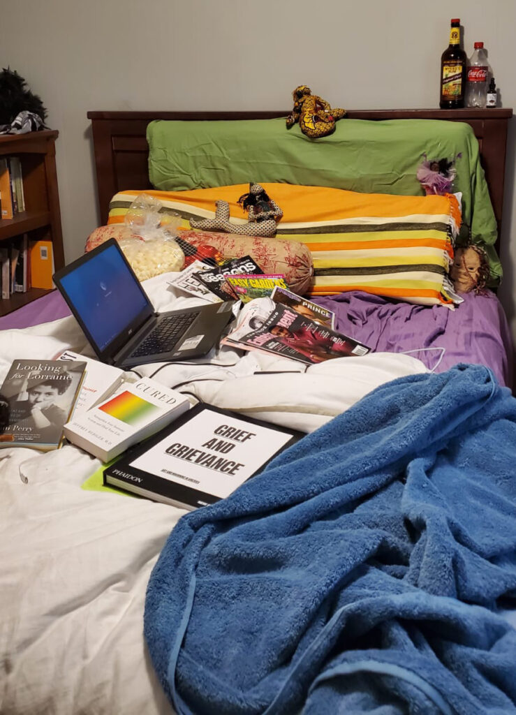 Bed with white duvet, blue towel, purple sheets, green and orange striped pillows, magazines, laptop, and many books.