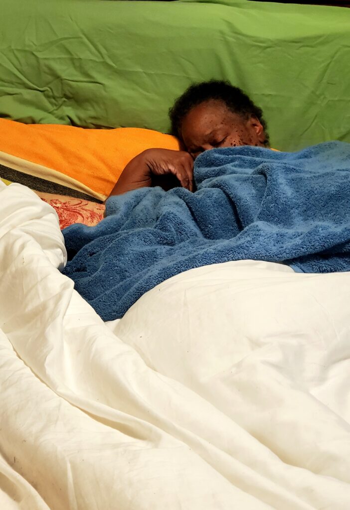 Person with dark skin lays in bed with eyes closed, face covered by blue towel.