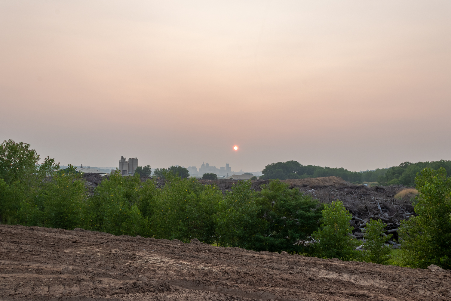 Sun sets over city skyline in hazy pink sky, behind brown dirt with tire tracks and line of green trees.