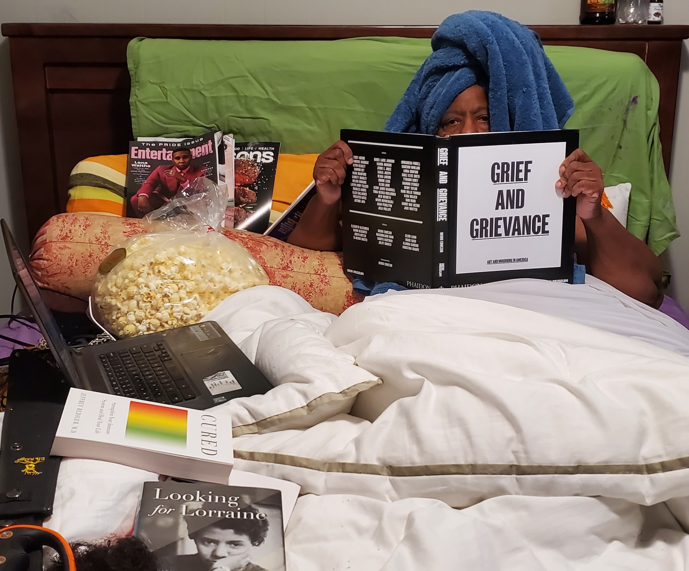 Person with dark skin and head wrapped in blue towel sits in bed full of books, holding up a book whose cover reads "GRIEF AND GRIEVANCE."