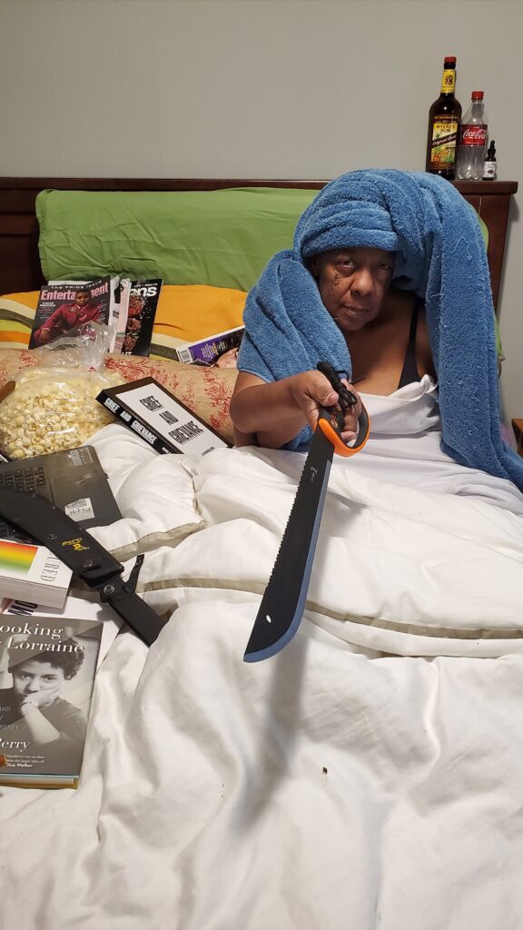 Person with dark skin and head wrapped in blue towel sits in bed covered with books, pointing a machete at the camera.