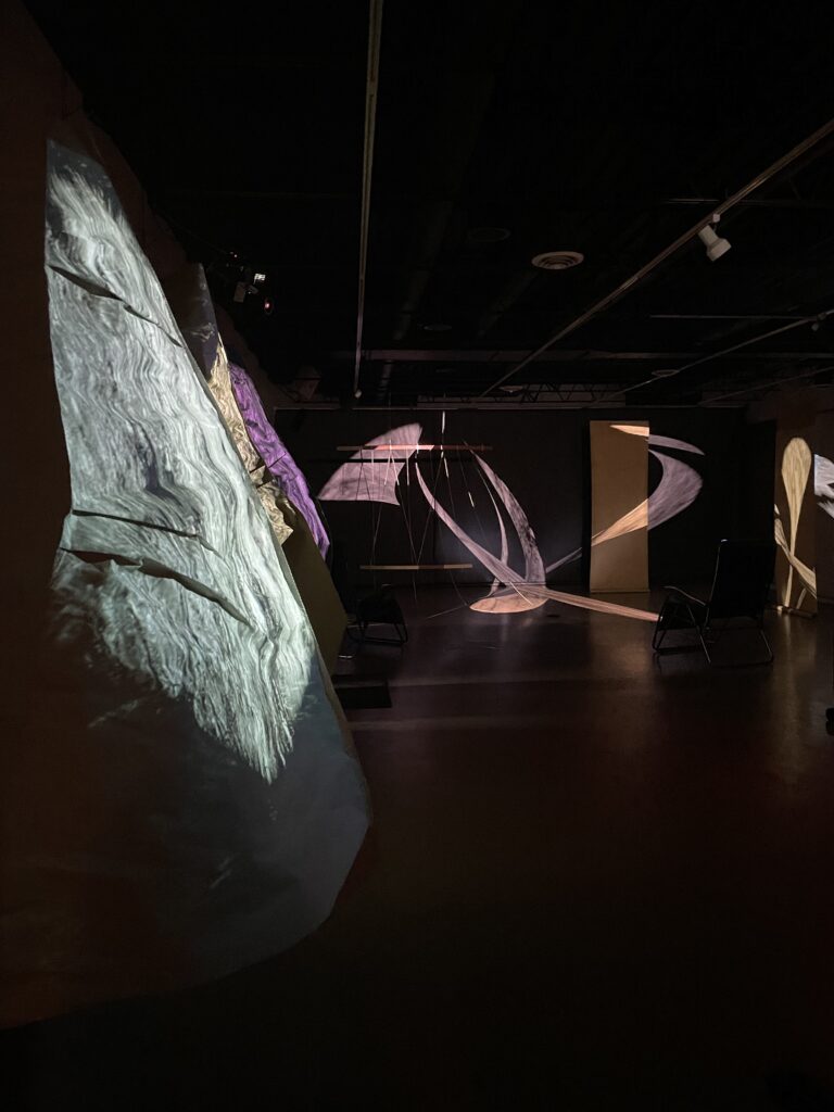 Dark gallery with projected patterns of light falling on walls and hanging sculptures.