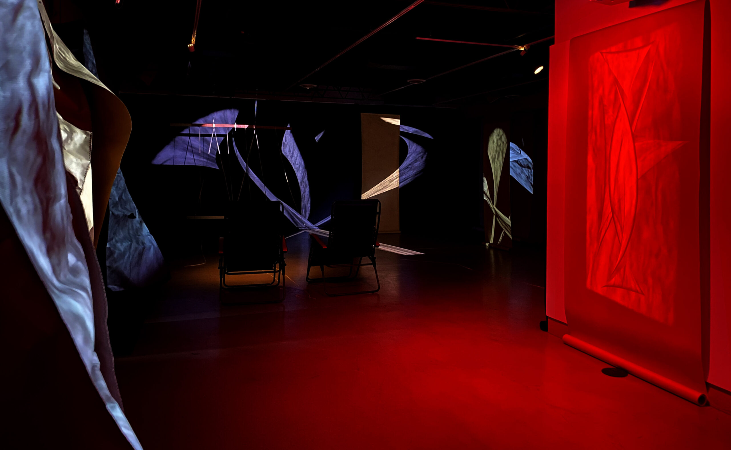 Dark room with projected pattern of purple and gold light, wash of red light on floor and wall, and two chairs in shadow.