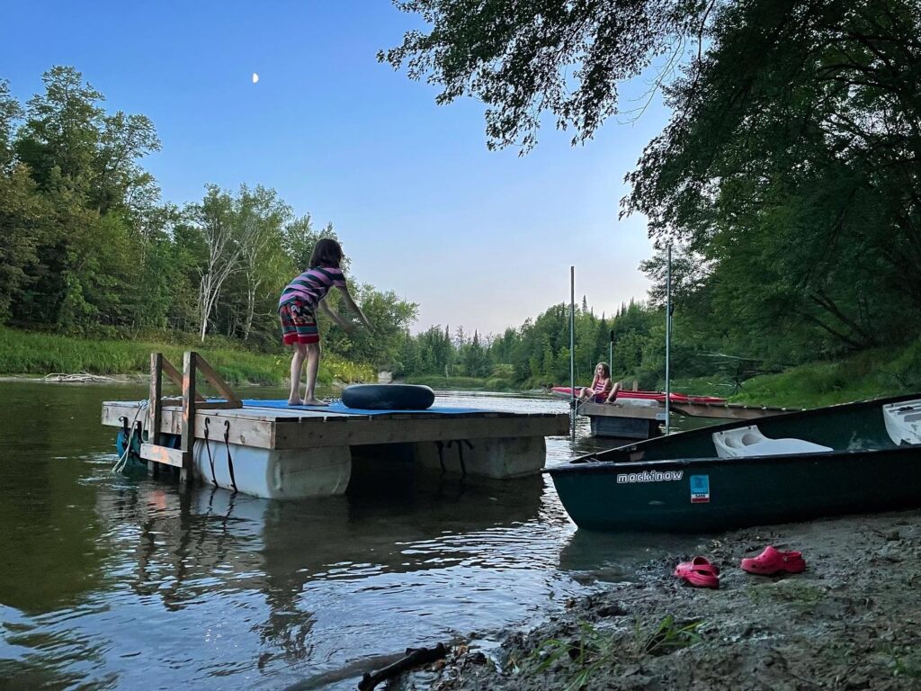 Two children on swimming platforms in low river, with dark green canoe on shoreline.