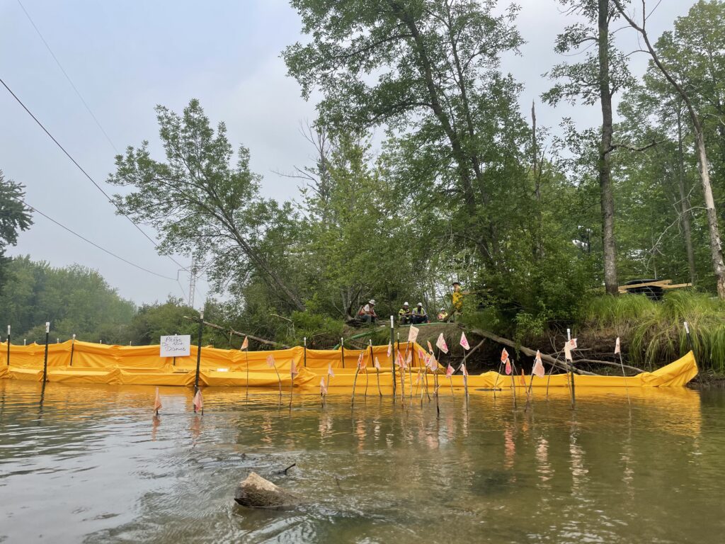 Yellow fencing and small flags in river, with green trees and group of people wearing hard hats in background.