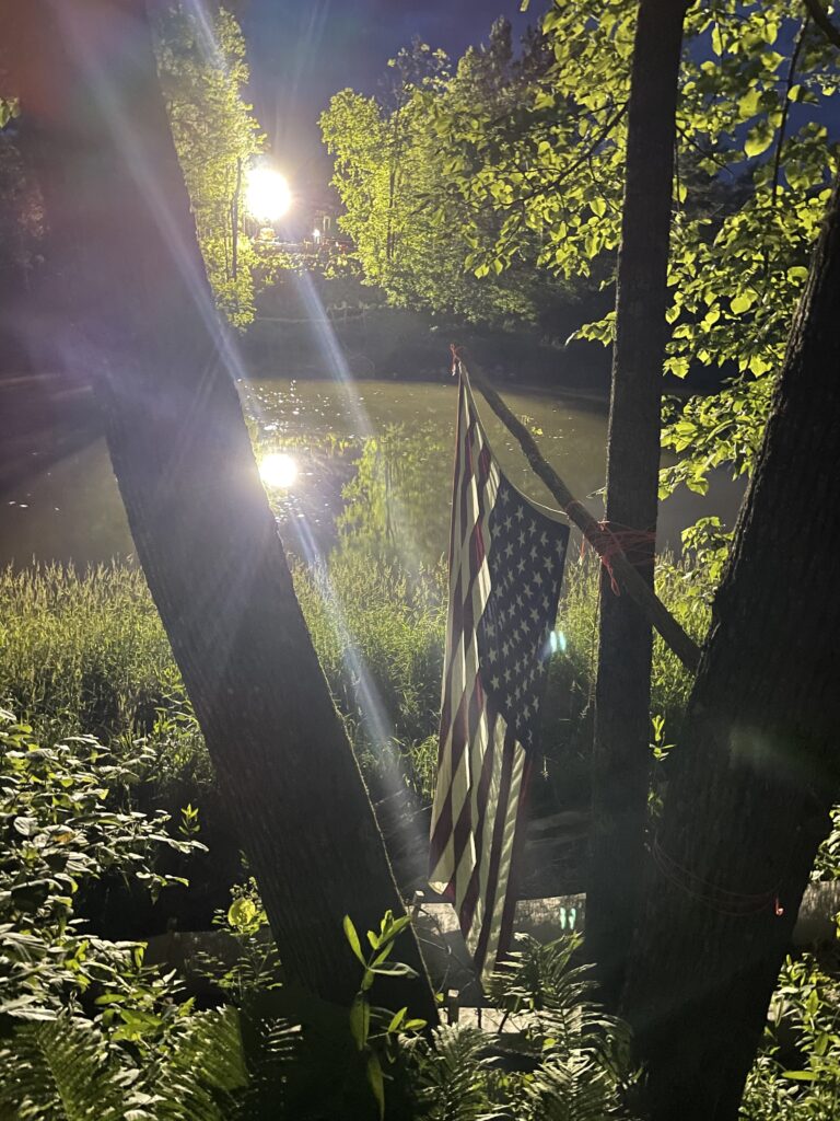United States flag hangs backwards over a river, with green trees and setting sun.
