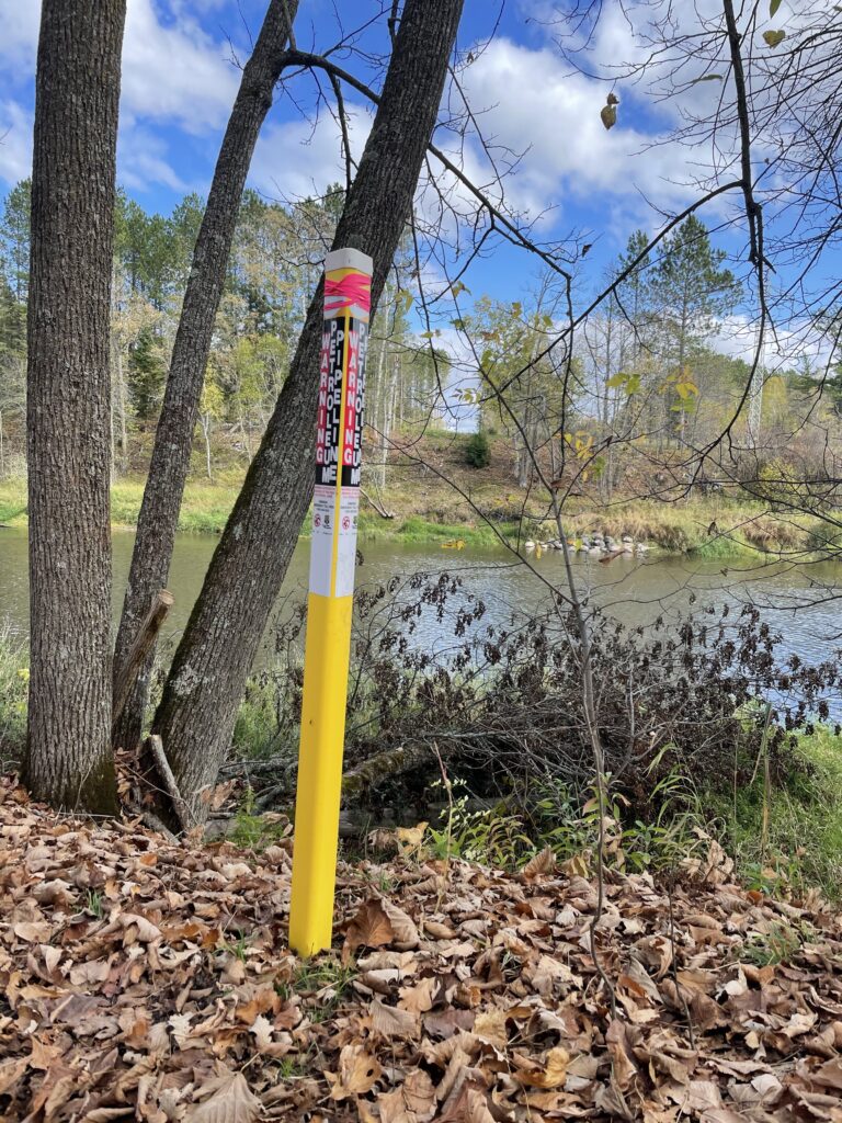 Yellow stake on riverbank reads: "WARNING / PETROLEUM / PIPELINE."