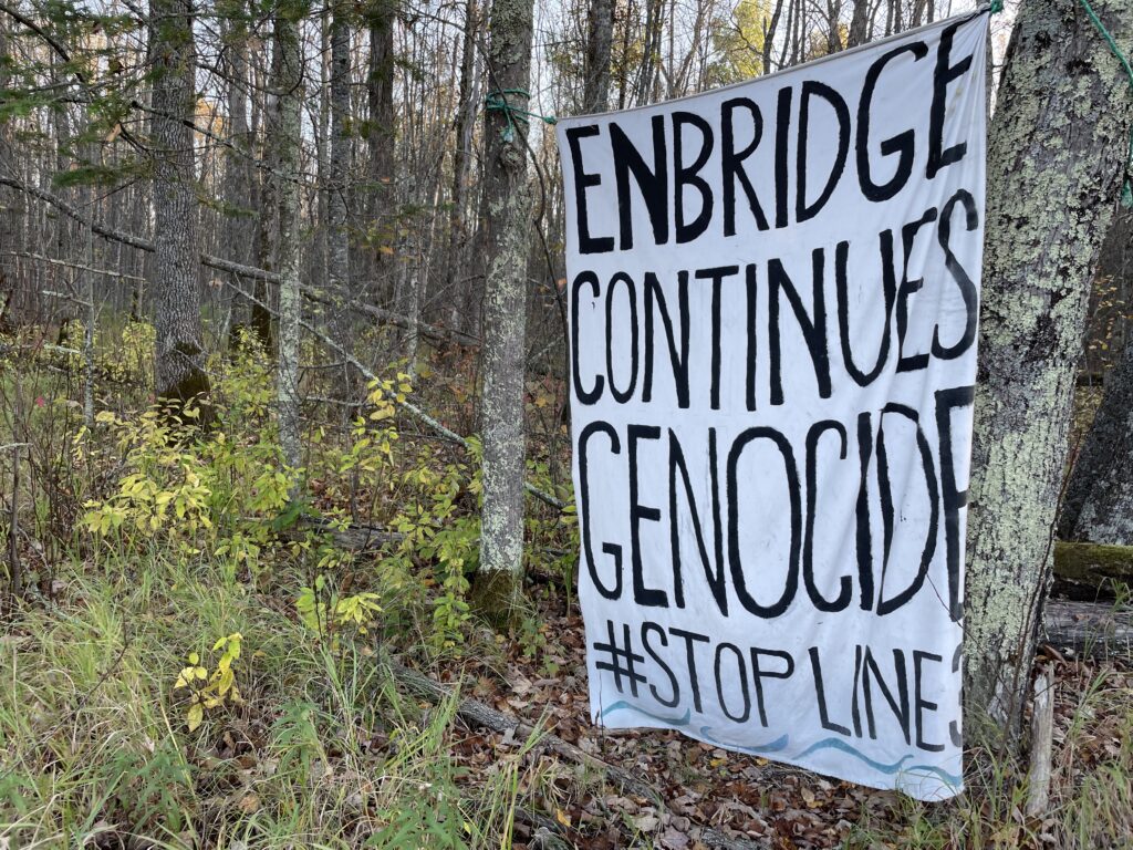 White sheet hangs in woods, painted with the words: "ENBRIDGE CONTINUES GENOCIDE #STOPLINE3."