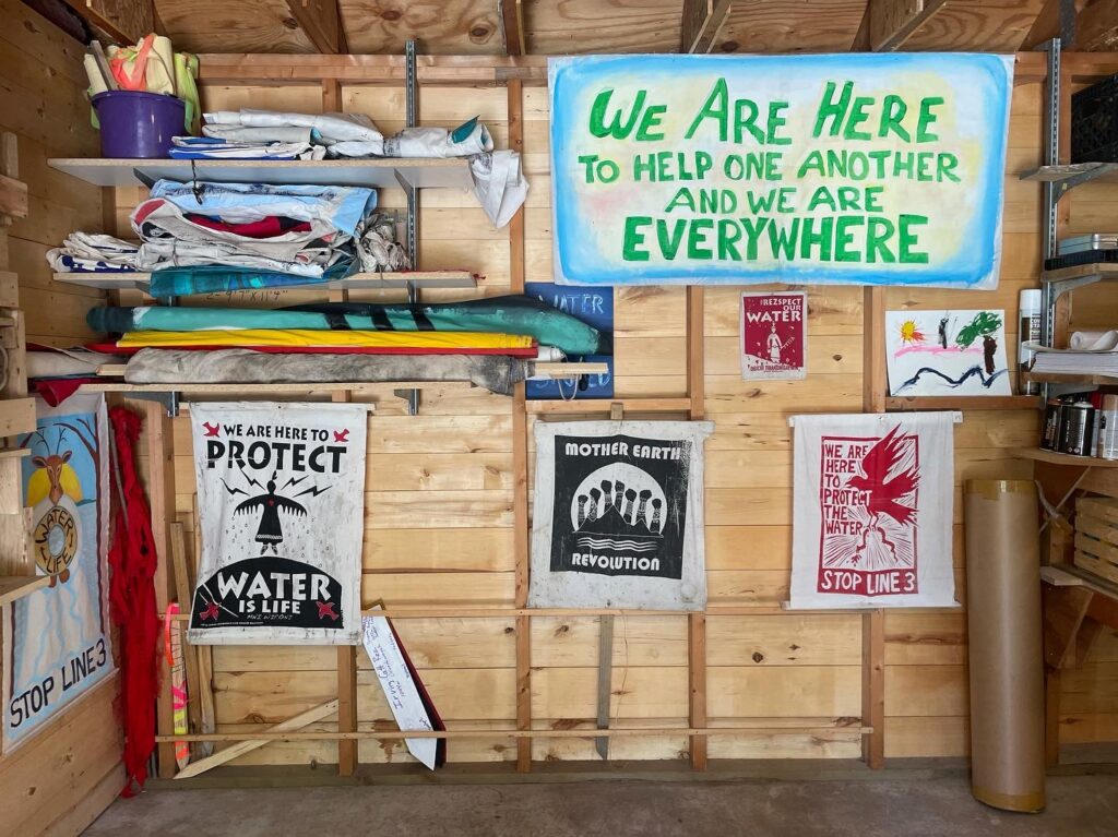 Art supplies and prints hang on wood wall, reading: "We are here to help one another and we are everywhere" / "stop line 3" / "we are here to protect, water is life" / "mother earth revolution."