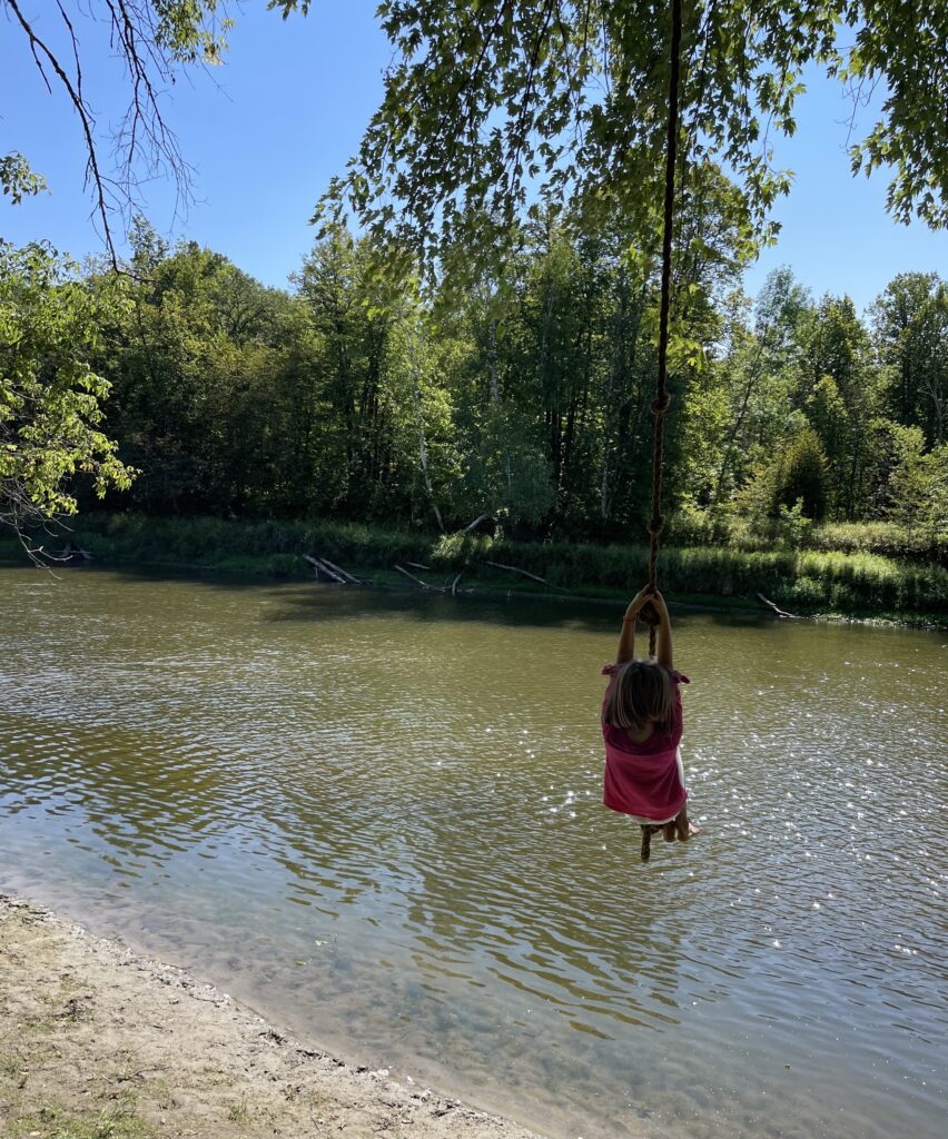 Child in pink shirt swings on rope swing over a river, with green trees and blue sky.