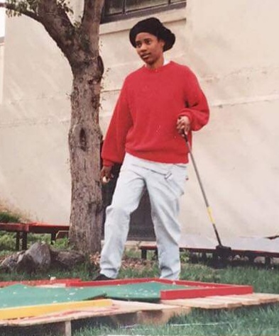 Black/biracial transmasculine nonbinary person wears black hat, red sweater, and khaki pants and plays mini golf.