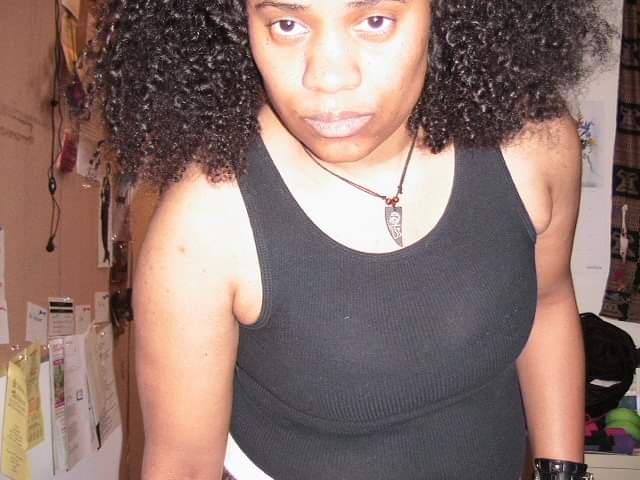 Black/biracial transmasculine nonbinary person wears black tank top and necklace, while looking at camera.