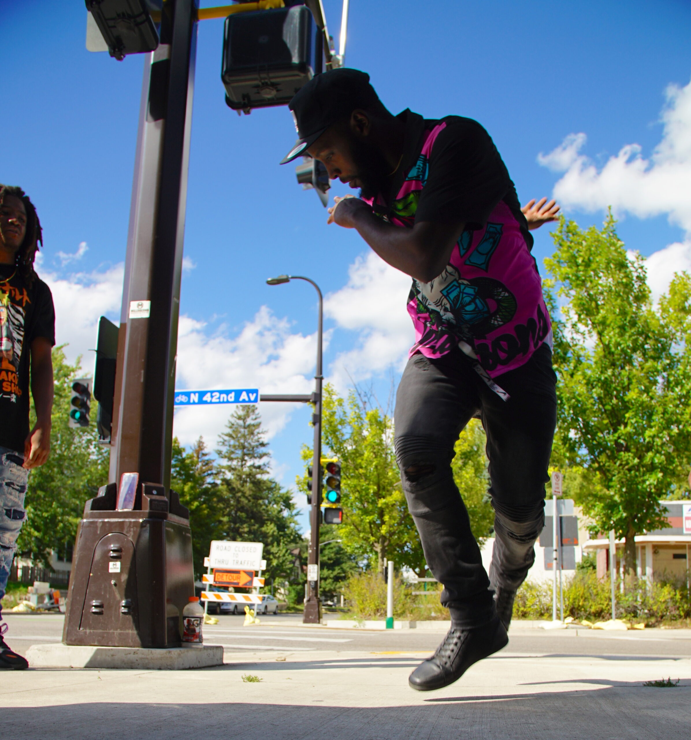 Person dancing on sidewalk, jumping and twisting over their right shoulder, with street signs, trees, and blue sky in background.