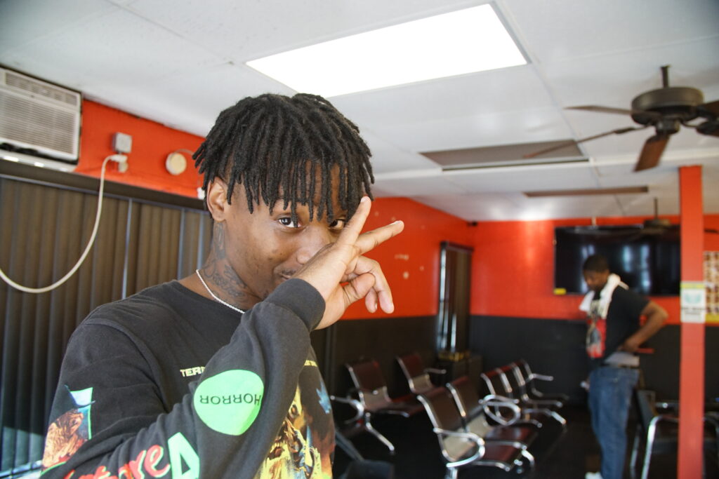 Person with dark skin and short locs gestures in front of their face and looks at camera, with interior of barber shop in background.