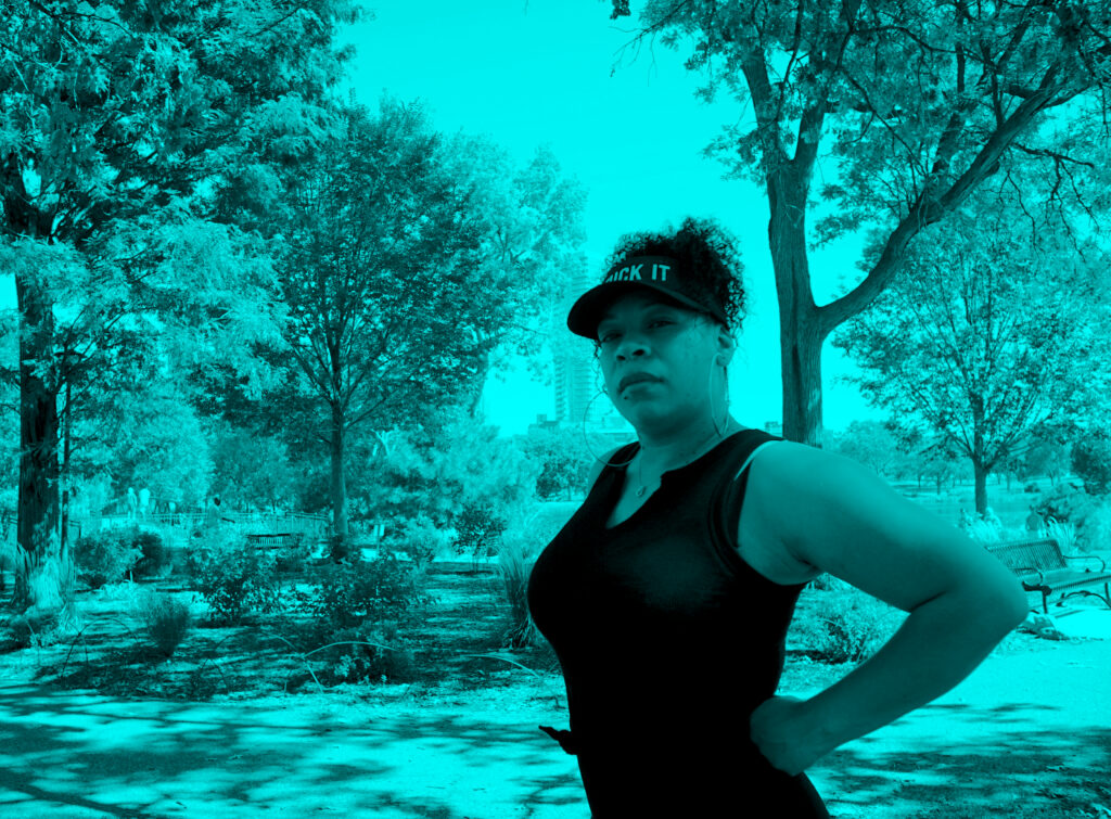 Person with black visor, ponytail, and black tank top looks at camera with hands on hips, in turquoise-tinted photo.