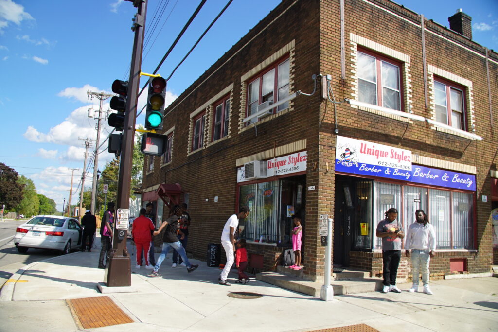 Group of people gather outside storefront at corner of brick building, with sign reading "Unique Stylez."