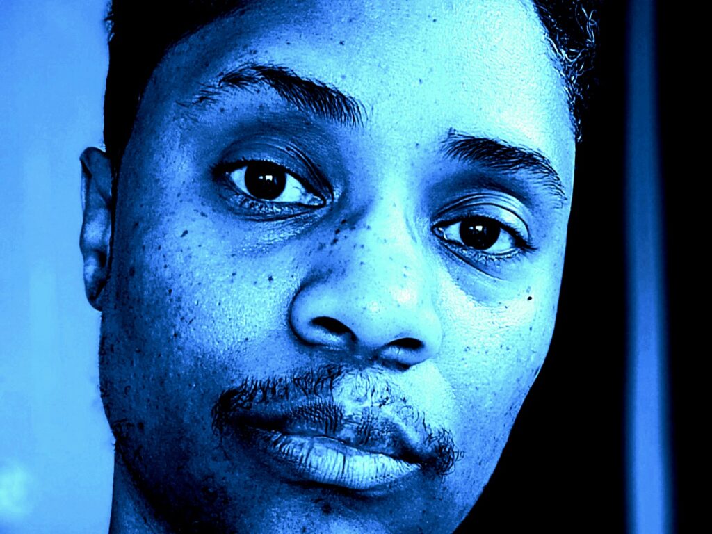 Blue-filtered photo of Black/biracial transmasculine nonbinary person with short hair and facial hair, looking into camera.