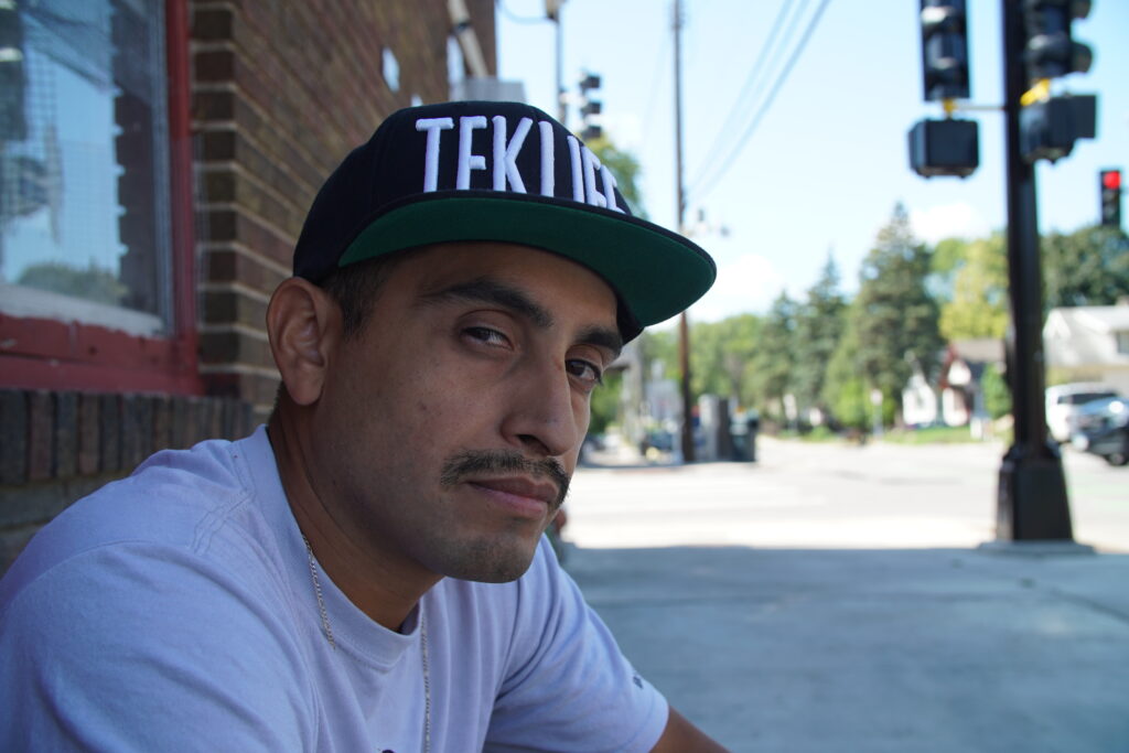 Person with medium skin and mustache, wearing white t-shirt and black hat, sits on street corner and looks at camera.