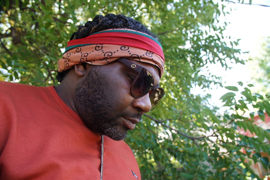 Person with dark skin, sunglasses, and red and orange headband looks down, with green tree in background.