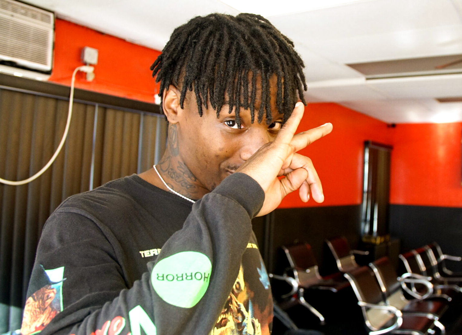 Person with dark skin and short locs gestures in front of their face and looks at camera, with interior of barber shop in background.