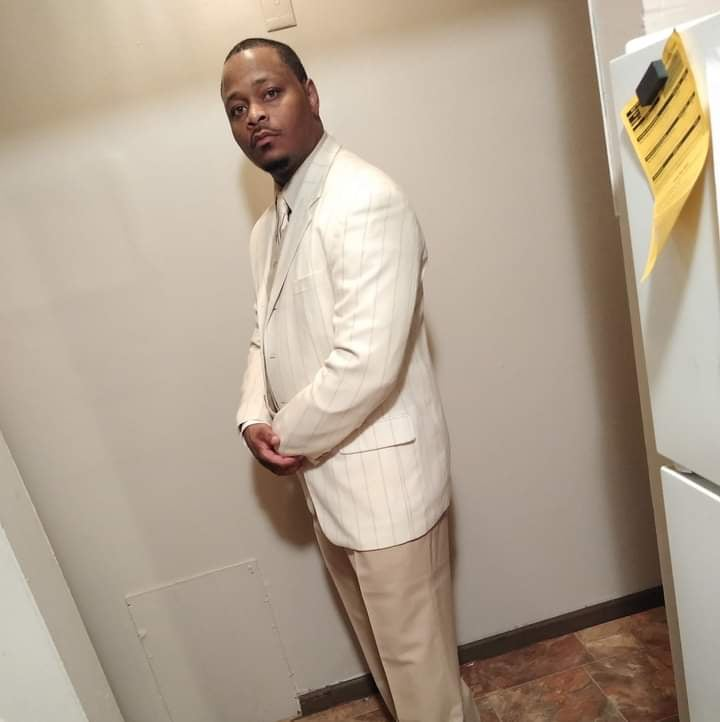 Person with dark skin wears white blazer, khaki pants, and stands facing side looking at camera.