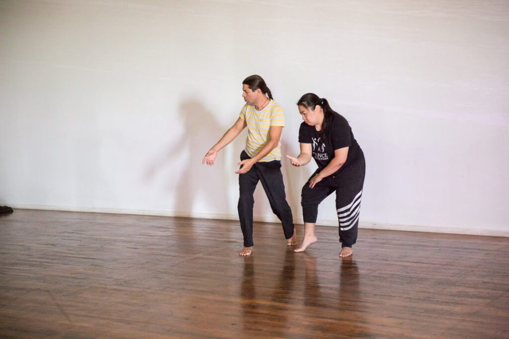 Two dancers standing close to each other soften their knees, spines, and arms, looking down and to the right, with wood floor and white wall surrounding them.