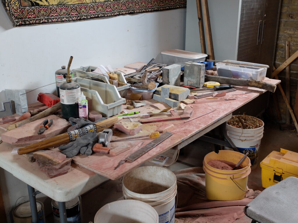 Studio table covered with stone, containers, tools, propane, with yellow and white buckets on floor.